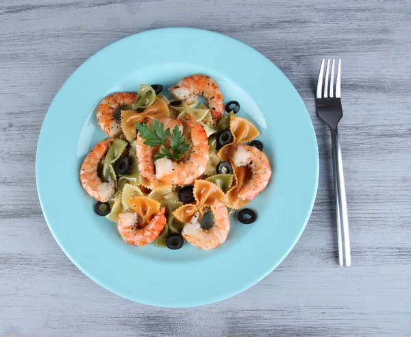 Fresh prawns with coloured macaroni, olives and parsley in a big blue round plate with a fork on wooden background — Stock Photo, Image