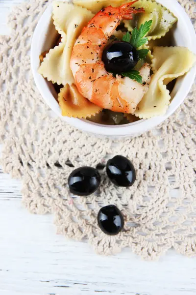 Fresh prawns with coloured macaroni and olives in a round bowl on a lace napkin on wooden background — Stock Photo, Image