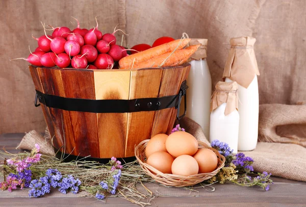 Gran canasta redonda con hierba seca, verduras, leche y huevos frescos sobre fondo de saco — Foto de Stock