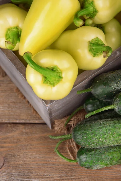 Gele paprika's in krat met komkommers op houten achtergrond — Stockfoto