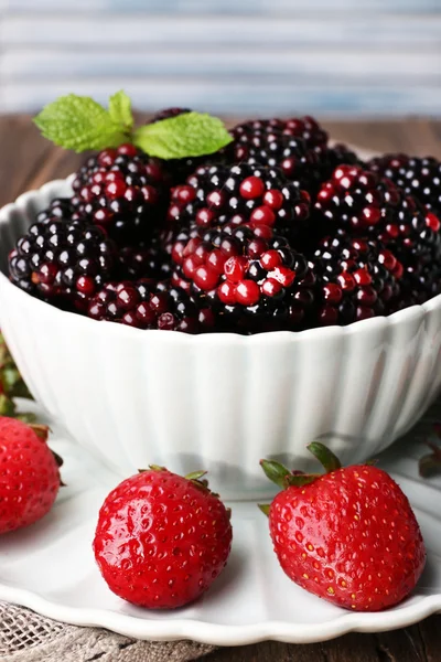 Bowl of blackberries and plate of strawberries on wooden table on light background — Stock Photo, Image