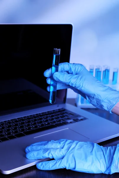 Cientista que introduz dados no computador portátil com tubos de ensaio num laboratório sobre fundo azul — Fotografia de Stock