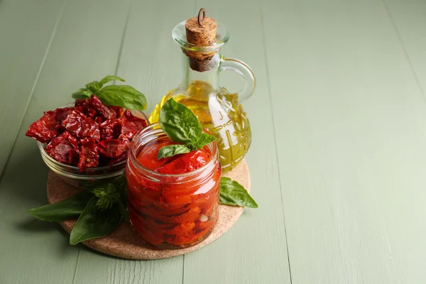 Tomates séchées au soleil dans un bocal en verre, huile d'olive en bouteille en verre, feuilles de basilic sur fond en bois de couleur — Photo