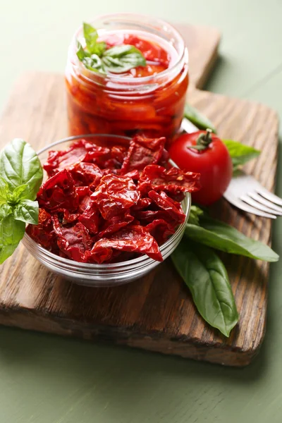 Tomates secados al sol en frasco de vidrio, hojas de albahaca en la tabla de cortar, sobre fondo de madera — Foto de Stock