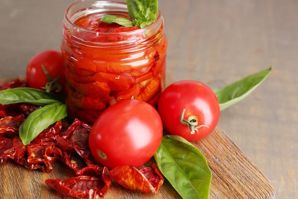 Sun dried tomatoes in glass jar, basil leaves on cutting board, on wooden background — Stock Photo, Image