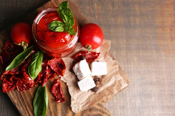 Sun dried tomatoes in glass jar, basil leaves and feta cheese on cutting board, on wooden background — Stock Photo, Image