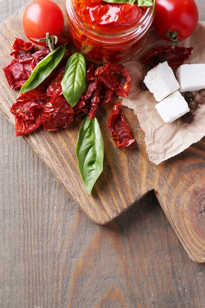 Sun dried tomatoes in glass jar, basil leaves and feta cheese on cutting board, on wooden background — Stock Photo, Image