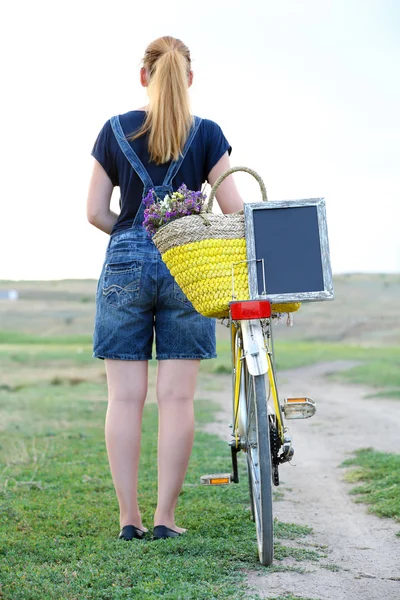 Femeie tânără cu bicicletă pe pajiște în timpul apusului de soare — Fotografie, imagine de stoc