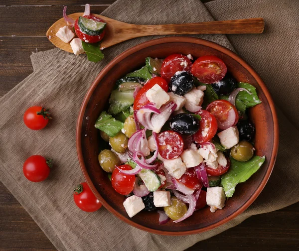 Greek salad served in brown bowl with bread on sacking napkin on wooden background — Stock Photo, Image