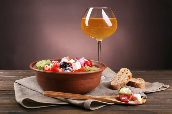 Salada grega servida em tigela marrom com fatia de pão e copo de vinho em guardanapo em mesa de madeira no fundo escuro — Fotografia de Stock