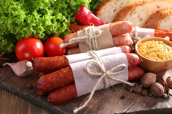 Assortimento di salsicce sottili, pane, senape in ciotola e spezie su tagliere, su fondo di legno — Foto Stock