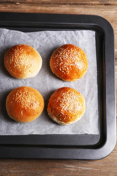 Tasty buns with sesame on oven-tray, on wooden background — Stock Photo, Image