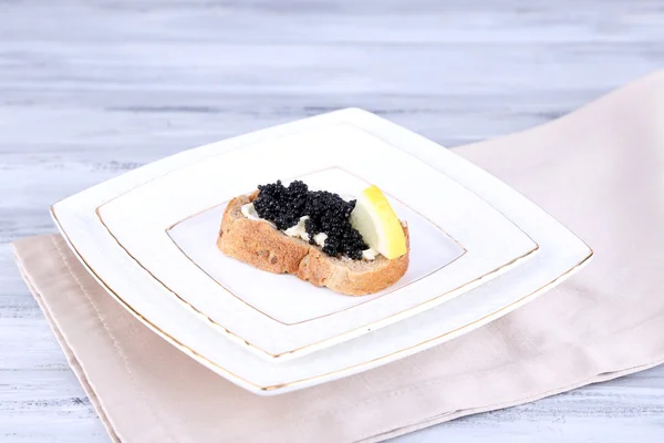 Slice of bread with butter, black caviar and lemon on plate on napkin on grey wooden background — Stock Photo, Image