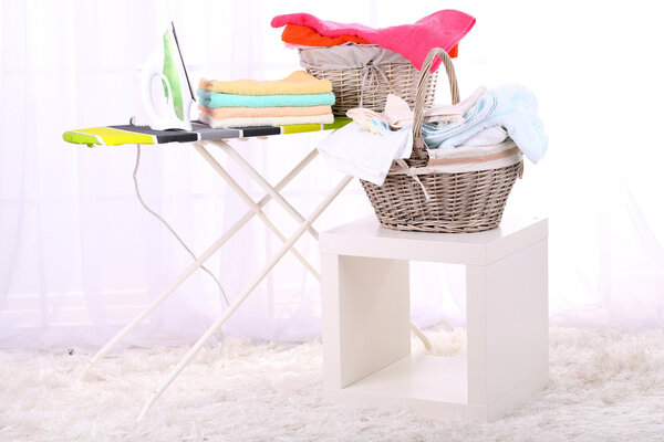 Basket with laundry and ironing board on light home interior background