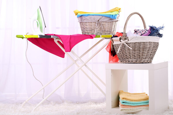 Baskets with laundry and ironing board on light home interior background