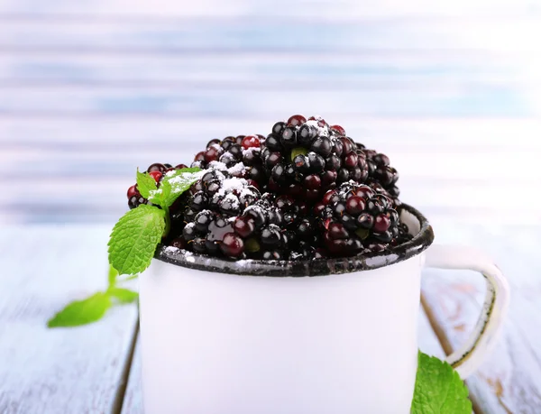 Metal mug of blackberries on wooden table on light background