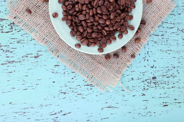 Granos de café en plato blanco sobre servilleta de saqueo sobre fondo de madera azul — Foto de Stock