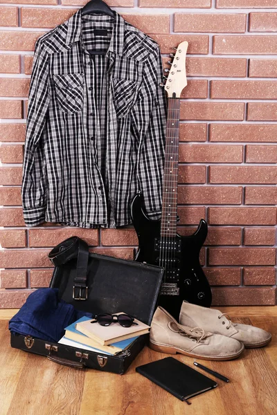 Guitar, clothes, boots and suitcase with books on floor on brick wall background — Stock Photo, Image