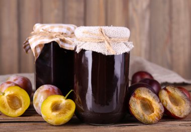 Tasty plum jam in jars and plums on wooden table on wooden background clipart