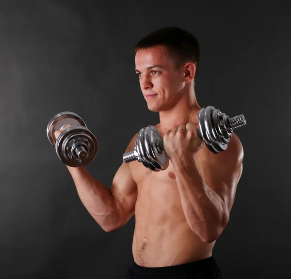 Handsome young muscular sportsman execute exercise with dumbbells on dark background — Stock Photo, Image