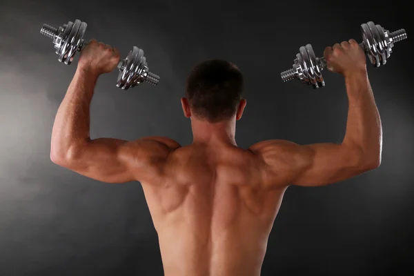 Handsome young muscular sportsman execute exercise with dumbbells on dark background — Stock Photo, Image