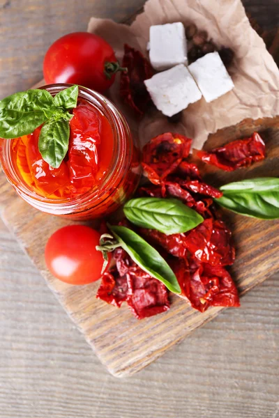 Solen torkade tomater i glasburk, basilikablad och fetaost på skärbräda, på trä bakgrund — Stockfoto