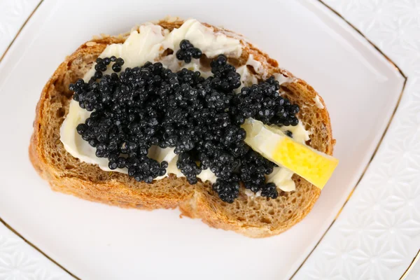 Slice of bread with butter, black caviar and lemon on plate closeup — Stock Photo, Image