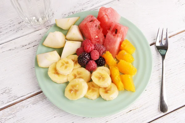 Sliced fruits and berries on plate with glass of water on wooden table — Stock Photo, Image