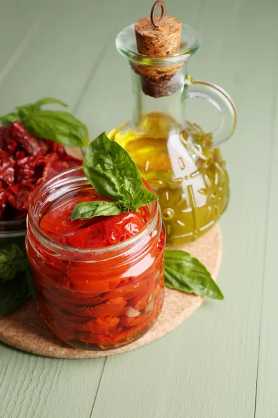 Tomates séchées au soleil dans un bocal en verre, huile d'olive en bouteille en verre, feuilles de basilic sur fond en bois de couleur — Photo