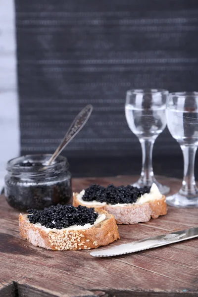 Slices of bread with butter and black caviar, glass jar of caviar and pair of small glasses on wooden table on dark background — Stock Photo, Image