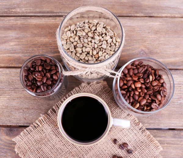 Mug of coffee and jars of coffee beans on sackcloth on wooden background — Stock Photo, Image