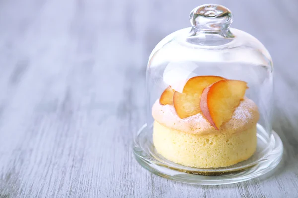 Вкусный мини-торт со свежим персиком на стеклянной тарелке с крышкой, на деревянном столе — стоковое фото