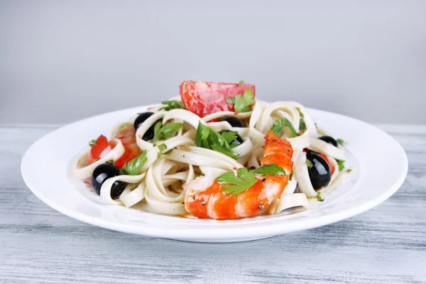 Gamberi freschi con spaghetti, olive, pomodori e prezzemolo in un grande piatto rotondo sul tavolo di legno su fondo grigio — Foto Stock