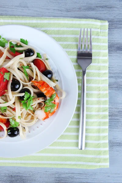 Gamberi freschi con spaghetti, olive, pomodori e prezzemolo in un grande piatto rotondo su tovagliolo su fondo di legno — Foto Stock