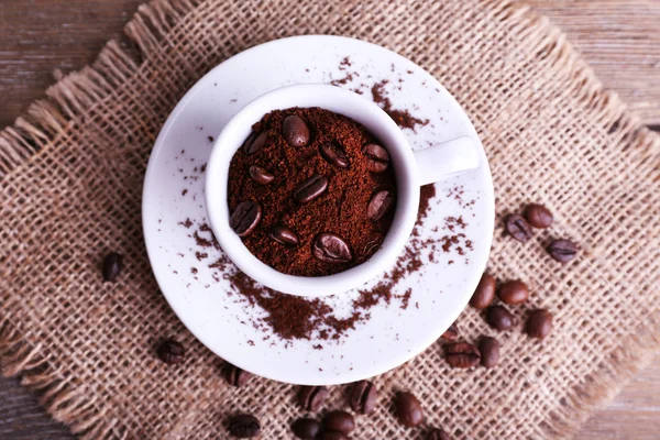 Mug of coffee beans and ground coffee on sackcloth on wooden background — Stock Photo, Image