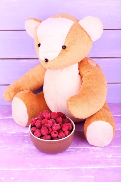 Toy bear and bowl of raspberries on wooden table on wooden wall background — Stock Photo, Image