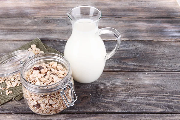 Granola fatta in casa in barattolo di vetro e latte fresco in brocca, su sfondo di legno a colori — Foto Stock