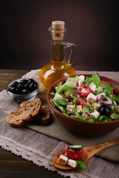 Bowl of Greek salad served with olive oil on sacking napkin on wooden table on dark background — Stock Photo, Image