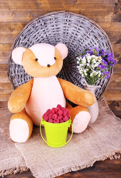 Toy bear, bucket of raspberries and vase of field flowers on sackcloth on wooden wall background — Stock Photo, Image