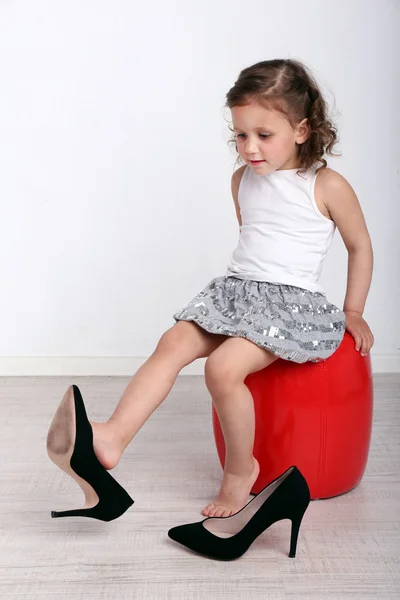 Beautiful small girl in big shoes sitting on leather ottoman on wall background