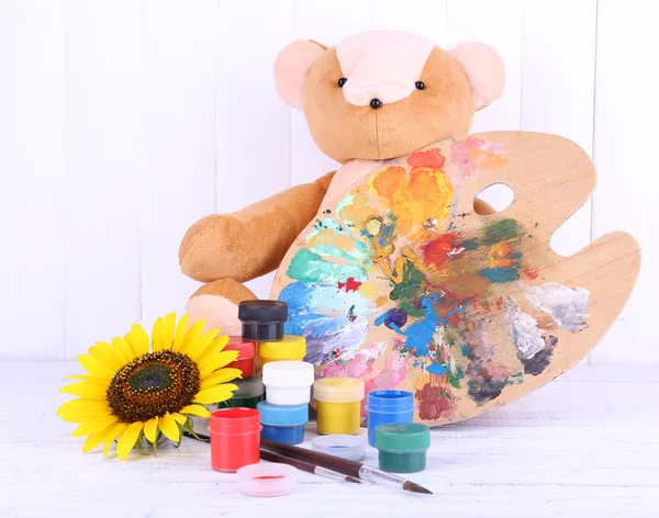 Toy bear, palette and paints on wooden wall background — Stock Photo, Image