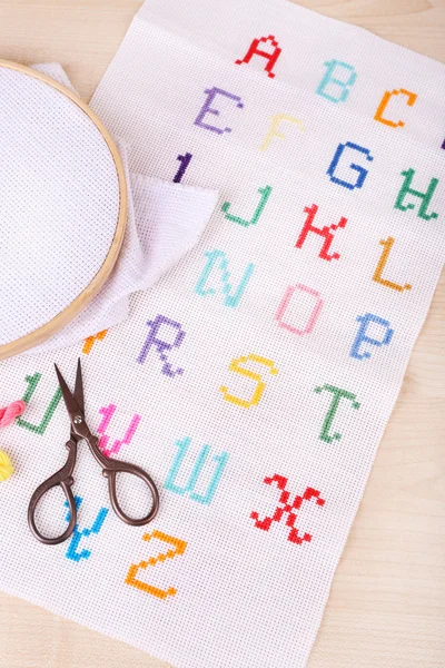 Handmade embroidered letters on white fabric and scissors on wooden background — Stock Photo, Image