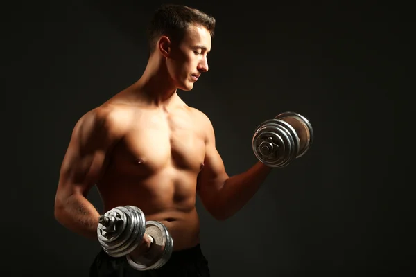 Handsome young muscular sportsman execute exercise with dumbbells on dark background — Stock Photo, Image