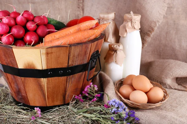 Stor rund korg med torkat gräs, grönsaker, mjölk och färska ägg på säckning bakgrund — Stockfoto