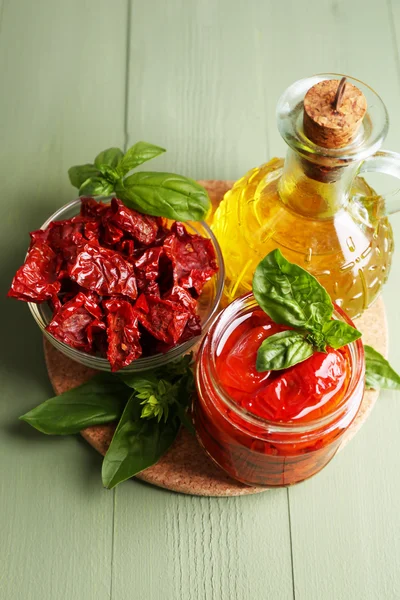 Tomates secados al sol en frasco de vidrio, aceite de oliva en botella de vidrio, hojas de albahaca sobre fondo de madera de color —  Fotos de Stock