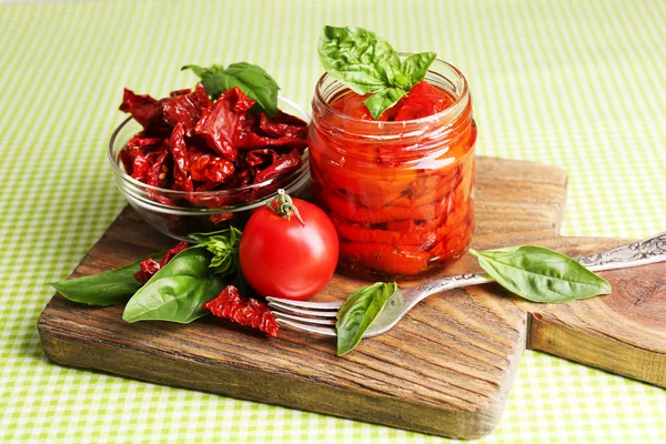 Sun dried tomatoes in glass jar, basil leaves on cutting board, on table background — Stock Photo, Image