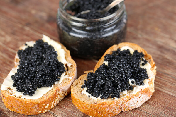 Slices of bread with butter and jar of black caviar on wooden background