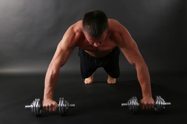 Handsome young muscular sportsman execute exercise with dumbbells on dark background — Stock Photo, Image