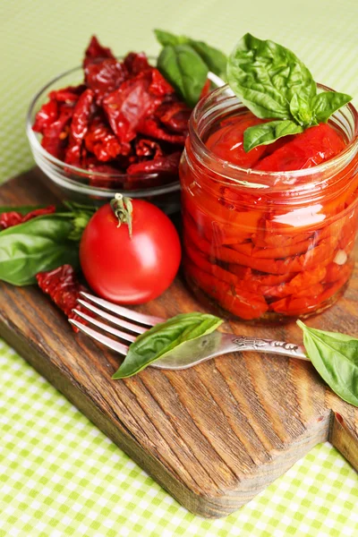 Sun dried tomatoes in glass jar, basil leaves on cutting board, on table background — Stock Photo, Image