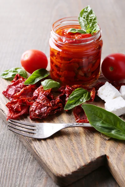 Tomates séchées au soleil dans un bocal en verre, feuilles de basilic et fromage feta sur planche à découper, sur fond en bois — Photo